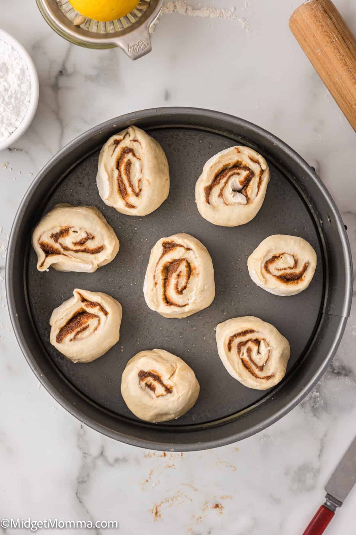 Unbaked cinnamon rolls arranged in a round baking pan on a marble surface with a rolling pin nearby.