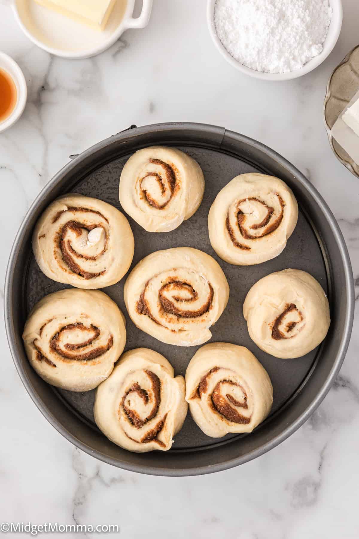 A round baking pan with eight uncooked cinnamon rolls on a marble countertop, surrounded by small bowls of ingredients like butter, powdered sugar, and vanilla extract.