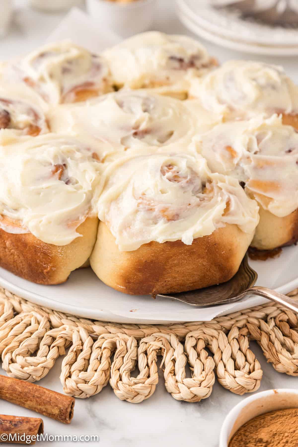 A plate of frosted cinnamon rolls on a woven placemat, with a bowl of cinnamon nearby.