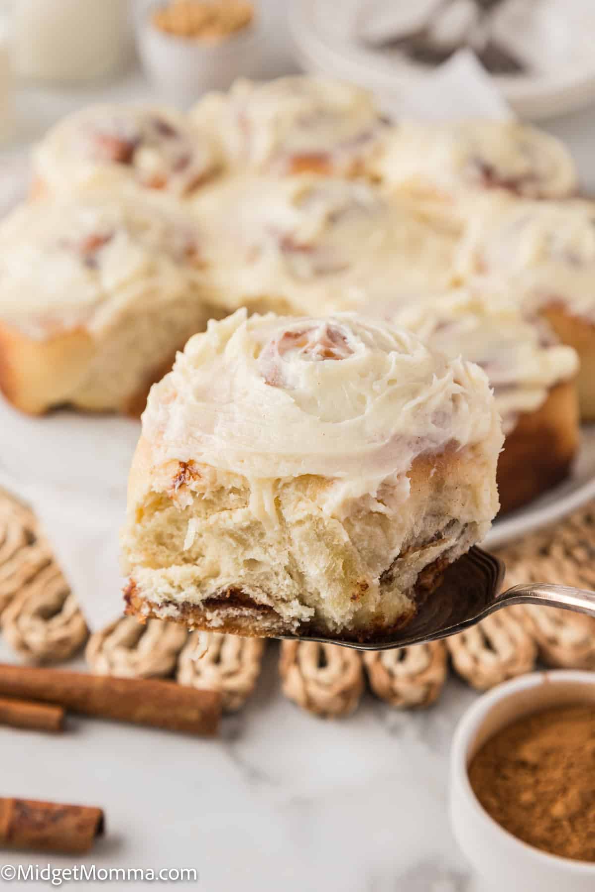 A close-up of a cinnamon roll with icing on a fork, with more cinnamon rolls blurred in the background.