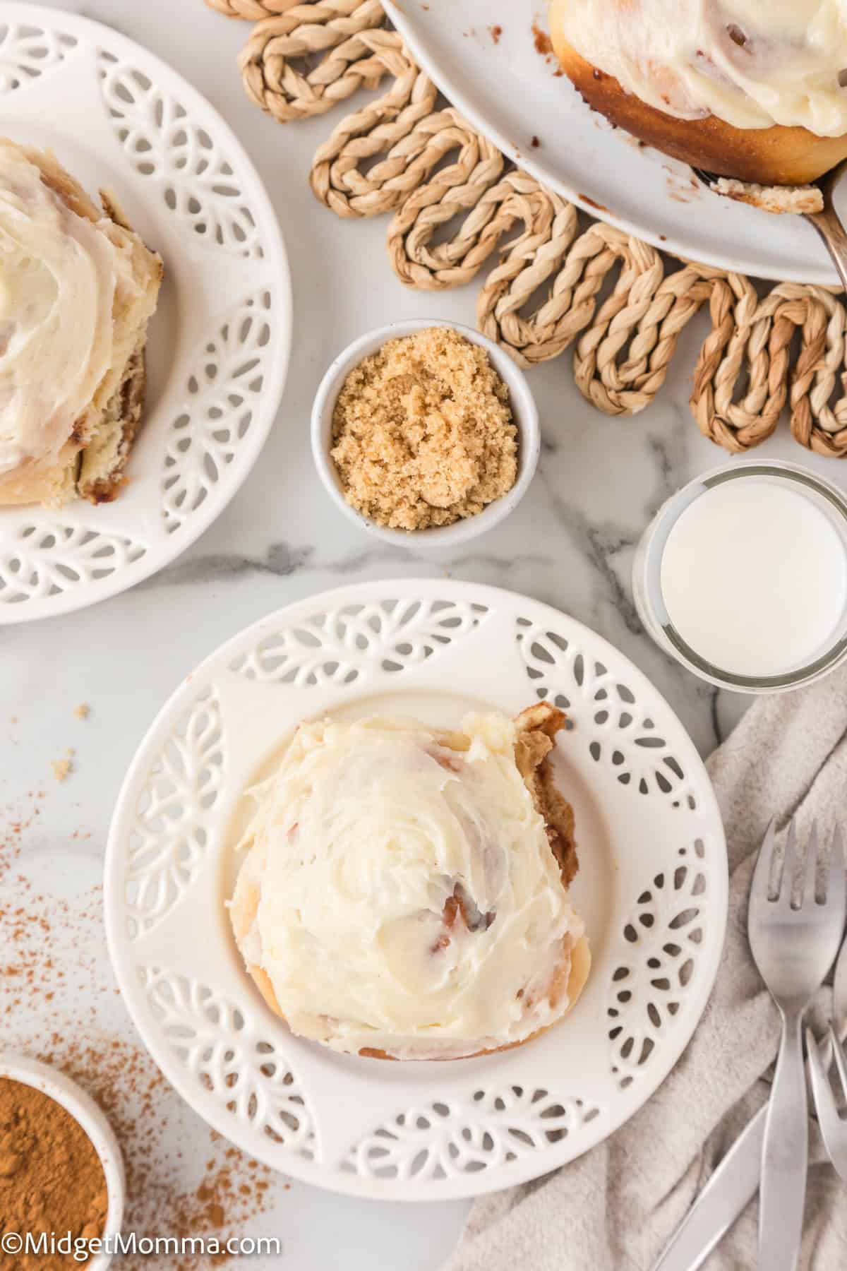 Frosted cinnamon rolls on white plates with small bowls of brown sugar and cinnamon, alongside a glass of milk and silverware on a white tablecloth.