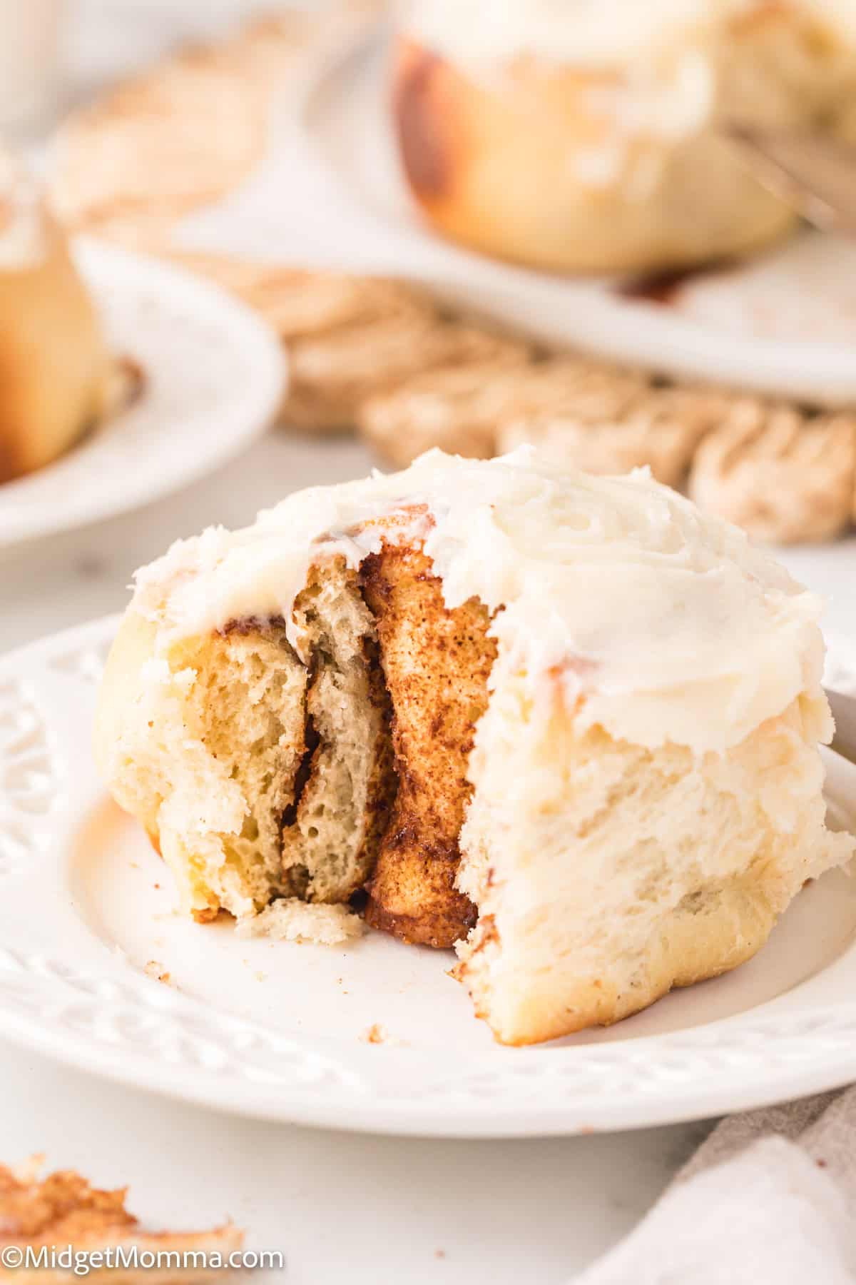 A frosted cinnamon roll is partially eaten on a white plate, showing layers of dough and cinnamon filling.