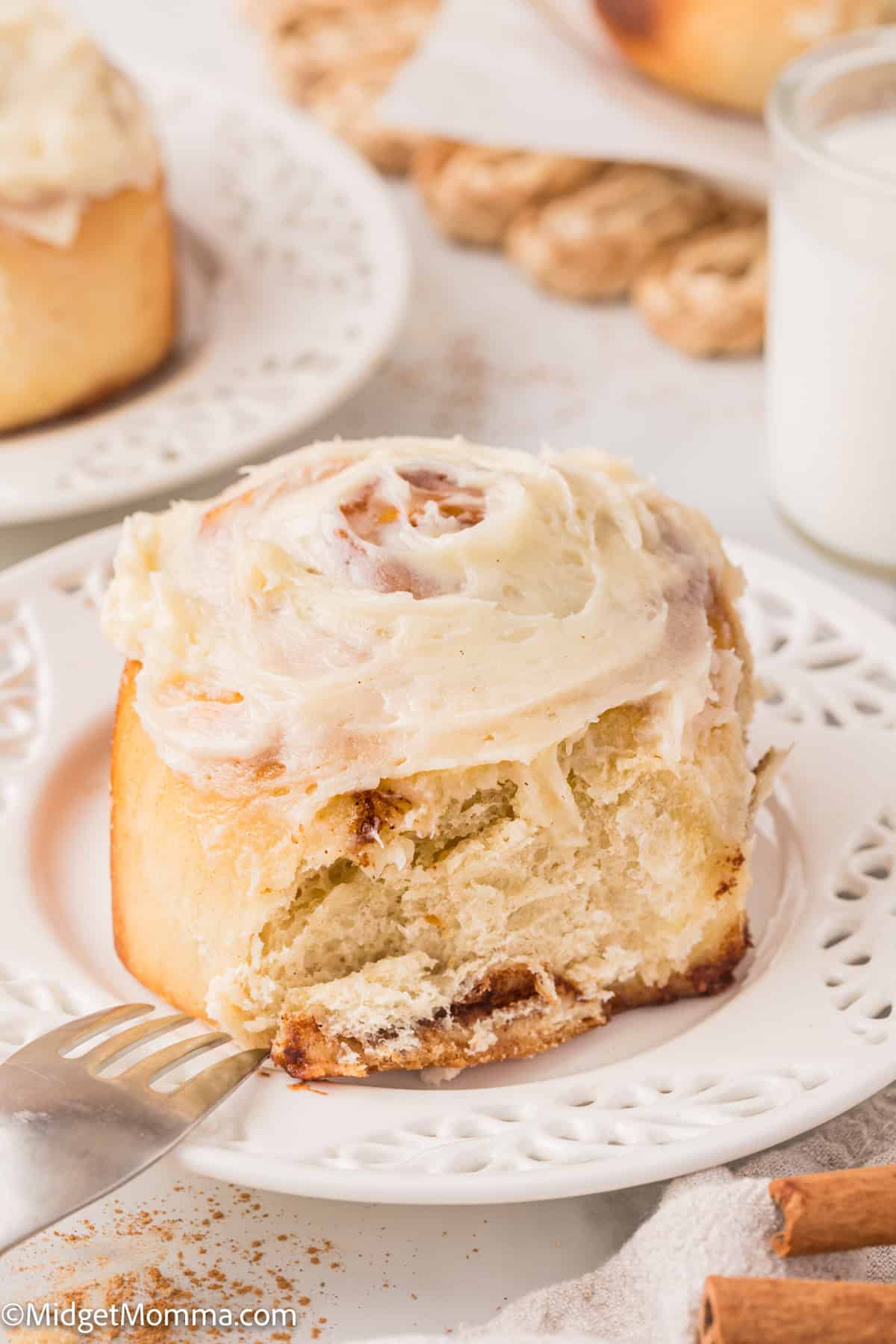 A frosted cinnamon roll on a white plate, partially eaten, with a fork beside it. A glass of milk and more cinnamon rolls are in the background.