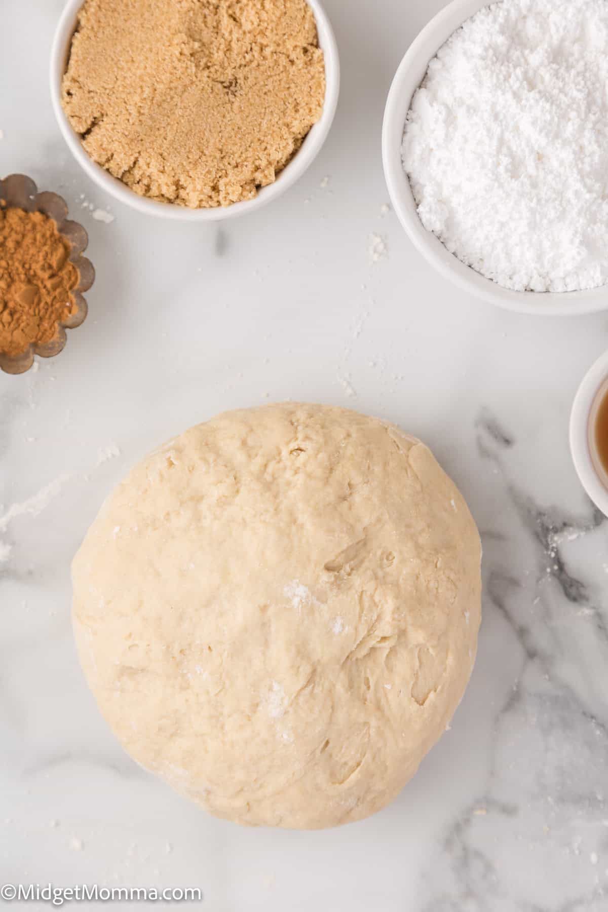 A ball of dough is surrounded by bowls of brown sugar, powdered sugar, cinnamon, and vanilla on a marble surface.