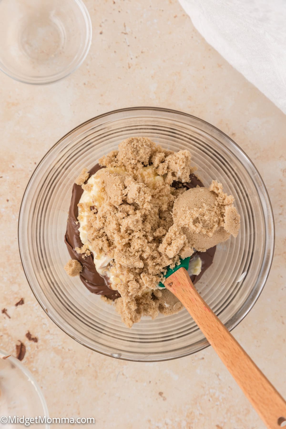 A glass bowl with brown sugar and chocolate, partially mixed with a wooden spatula, on a light countertop.