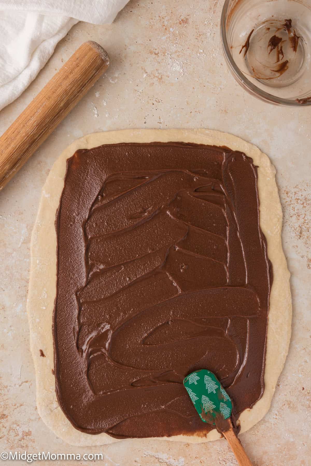 Dough rolled out with chocolate spread, a wooden rolling pin, and an empty glass bowl with remnants of chocolate nearby.