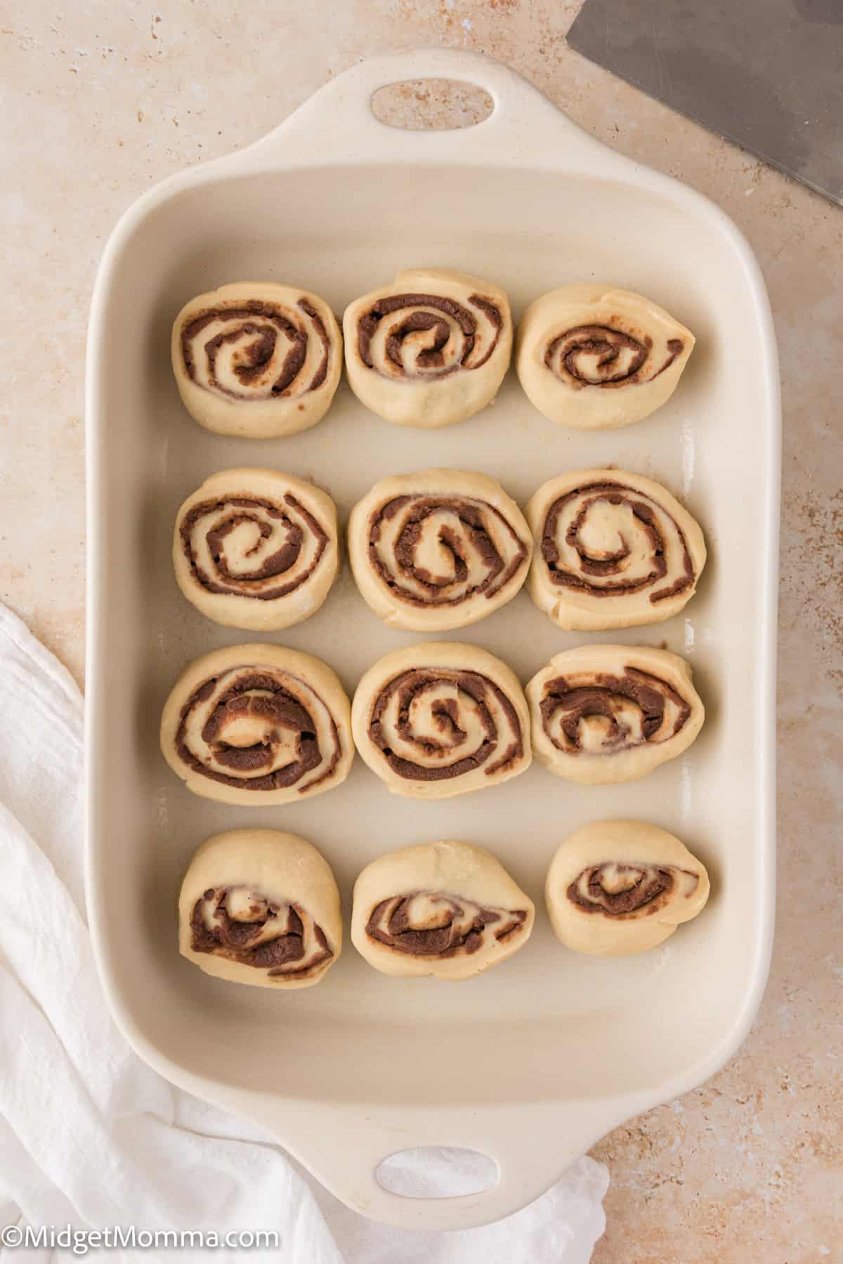 Baking dish with twelve unbaked cinnamon rolls arranged in three rows of four.