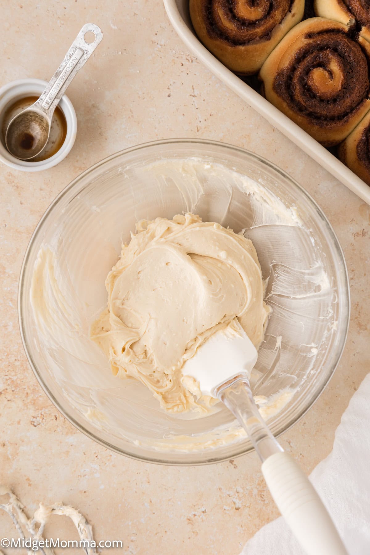 A bowl of creamy frosting with a spatula, next to a pan of cinnamon rolls and a tablespoon on a countertop.