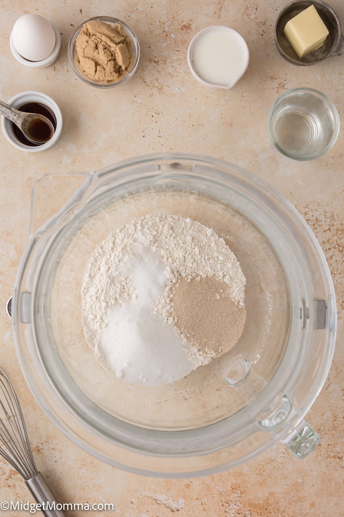 Top view of baking ingredients on a countertop: a bowl with flour, sugar, and yeast, surrounded by dishes of brown sugar, milk, butter, water, vanilla, an egg, and a whisk.