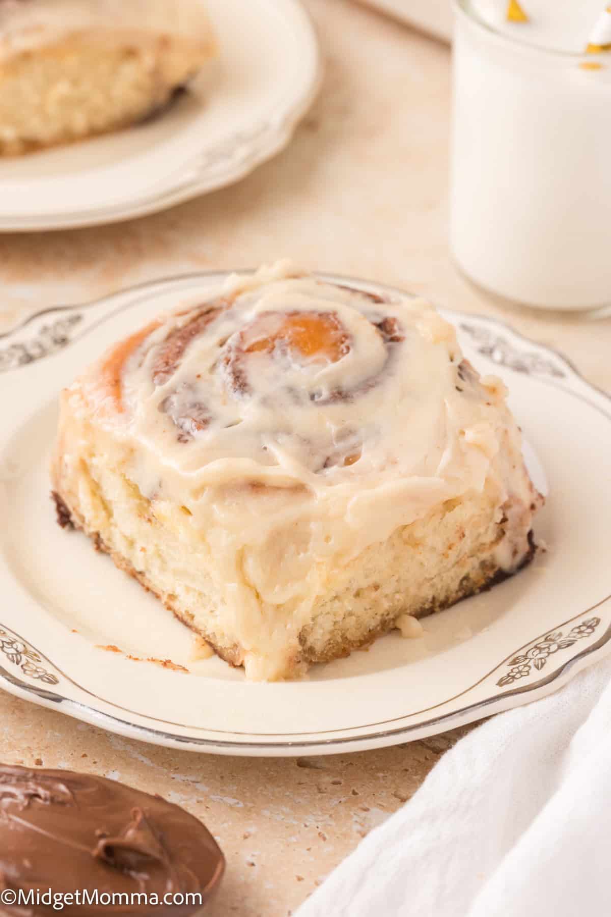 A frosted Nutella cinnamon roll on a decorative plate sits next to a glass of milk.