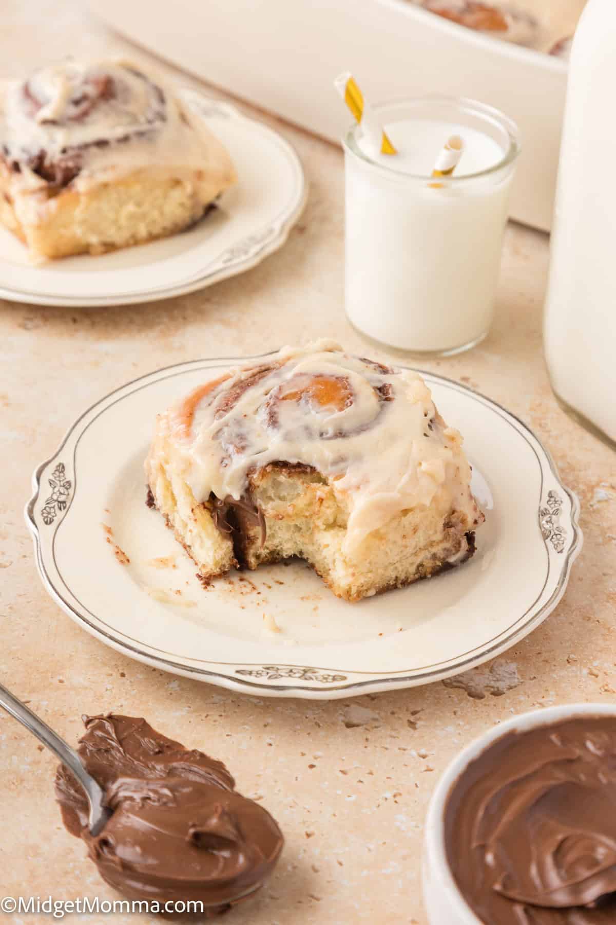 A nutella cinnamon roll with icing on a plate, a glass of milk, and a spoonful of chocolate spread nearby.