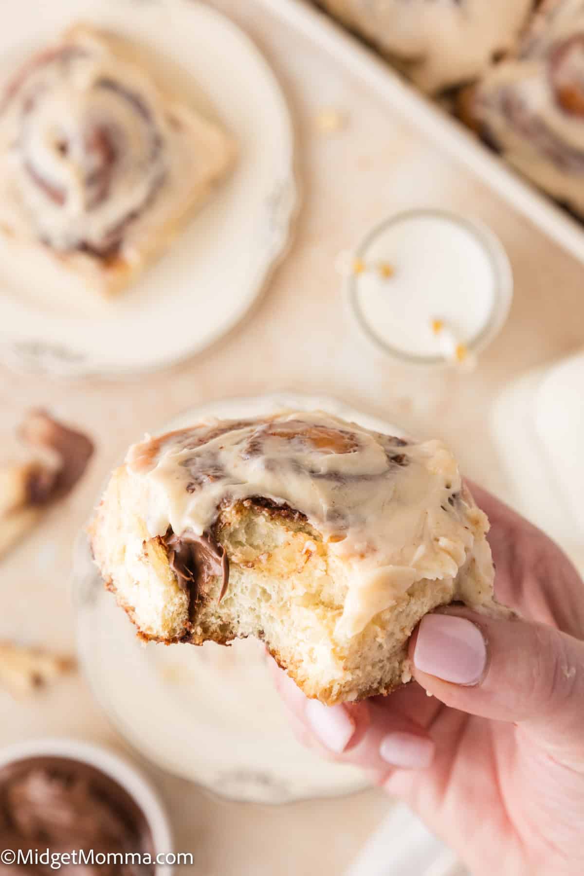 A hand holds a partially eaten cinnamon roll with icing. Additional cinnamon rolls and a small glass of milk are visible in the background.