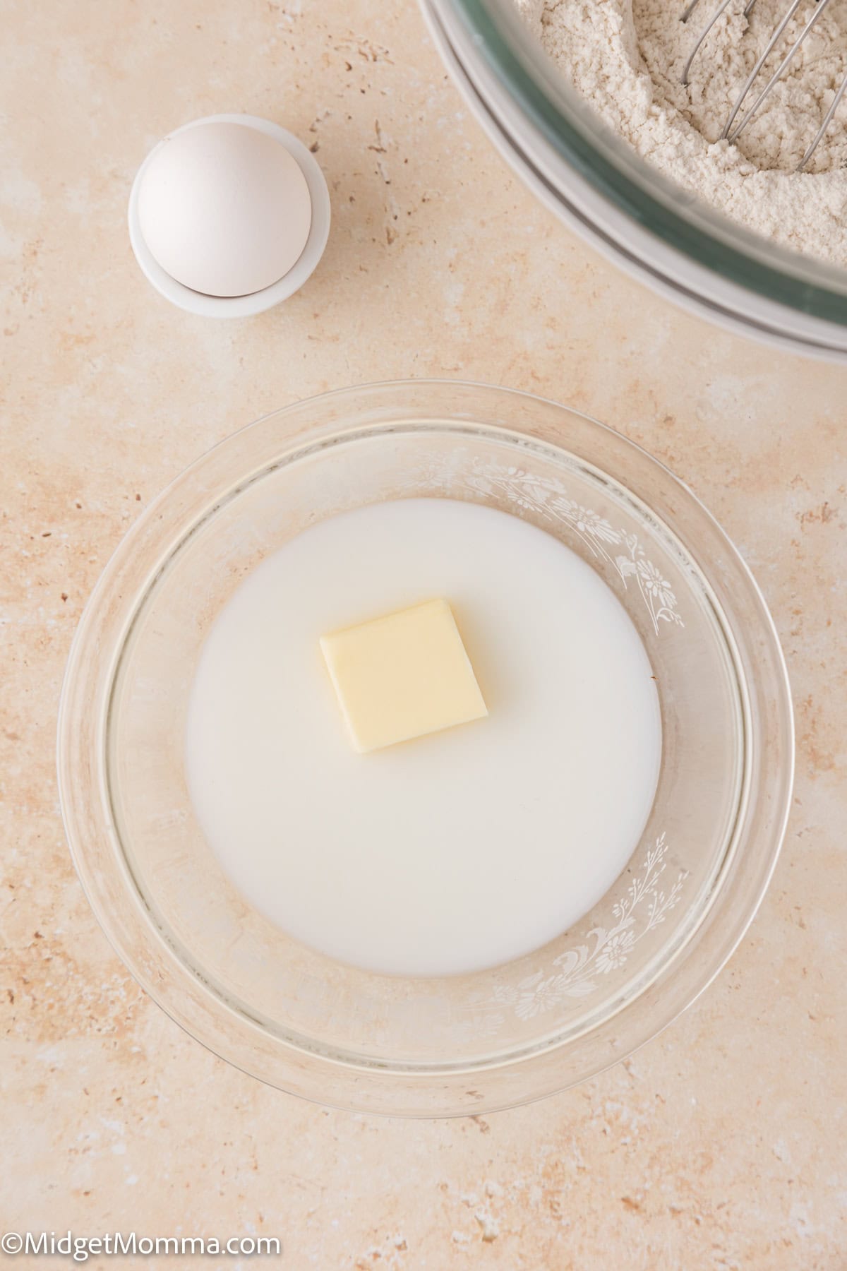 A stick of butter is placed in a bowl of milk. An egg rests in an egg cup, and flour is visible in a mixing bowl.