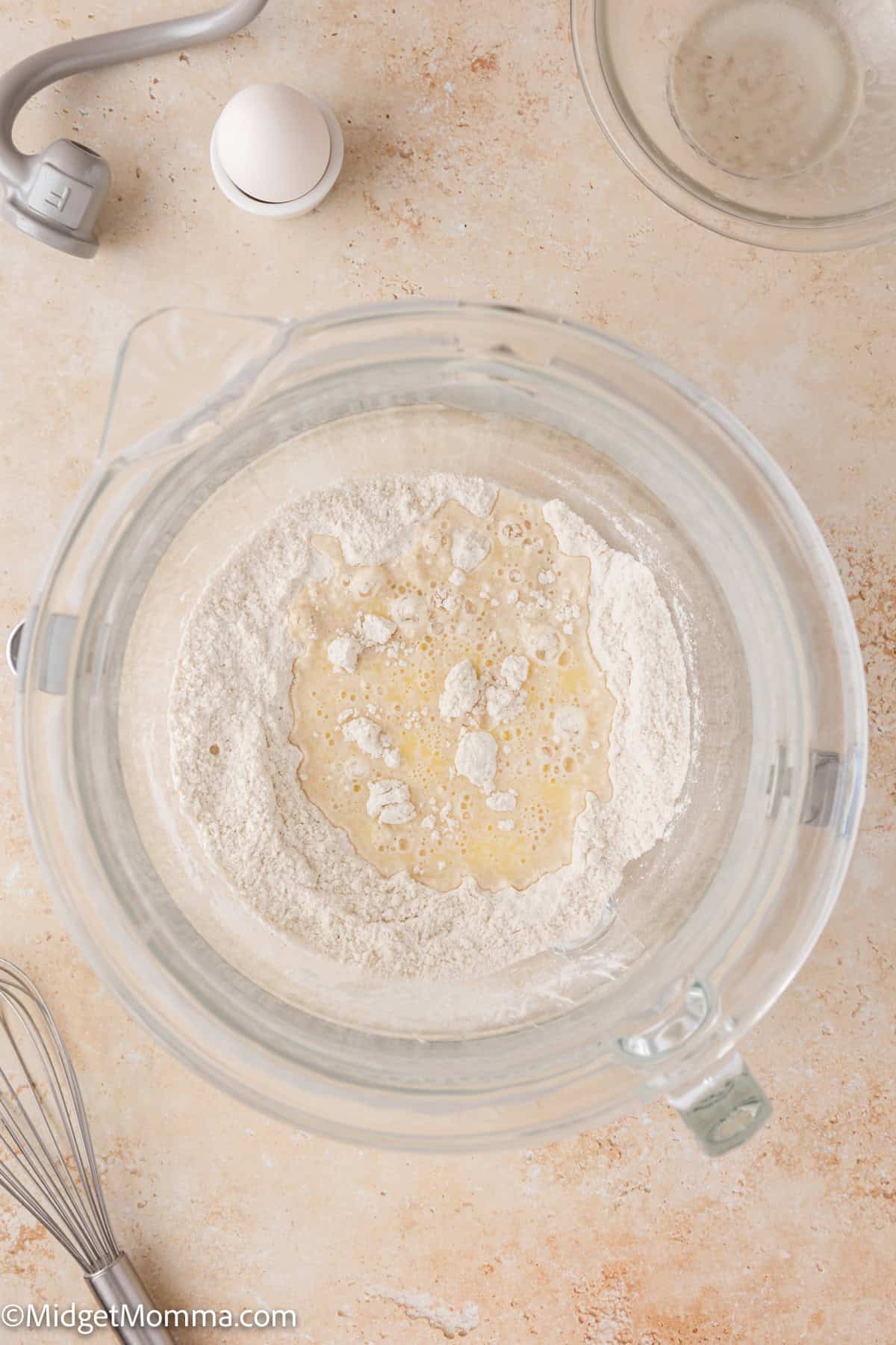 A mixing bowl with flour and wet ingredients on a table, surrounded by a whisk, cracked egg shell, and measuring cup.