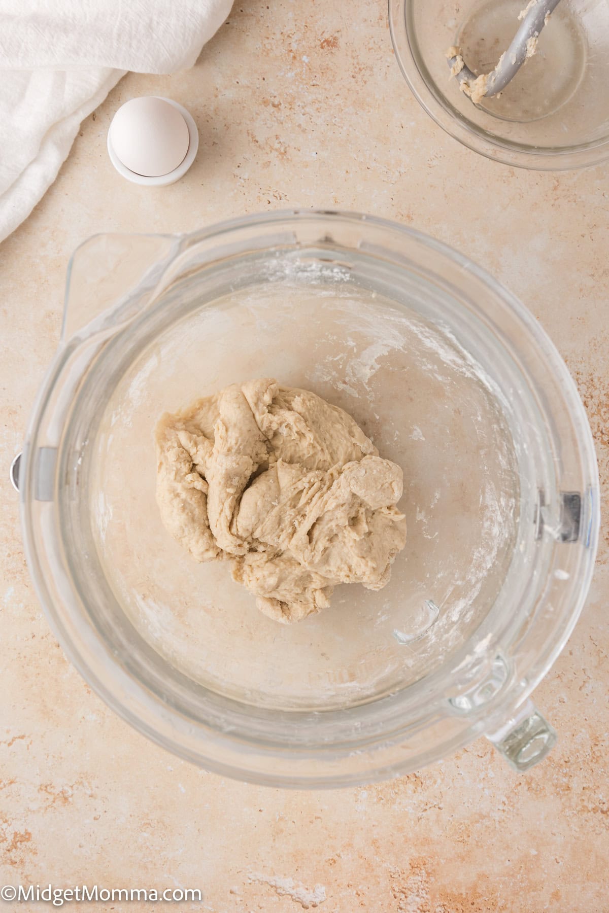 Ball of dough in a clear glass bowl on a light-colored surface, with a cracked egg and mixing attachment nearby.