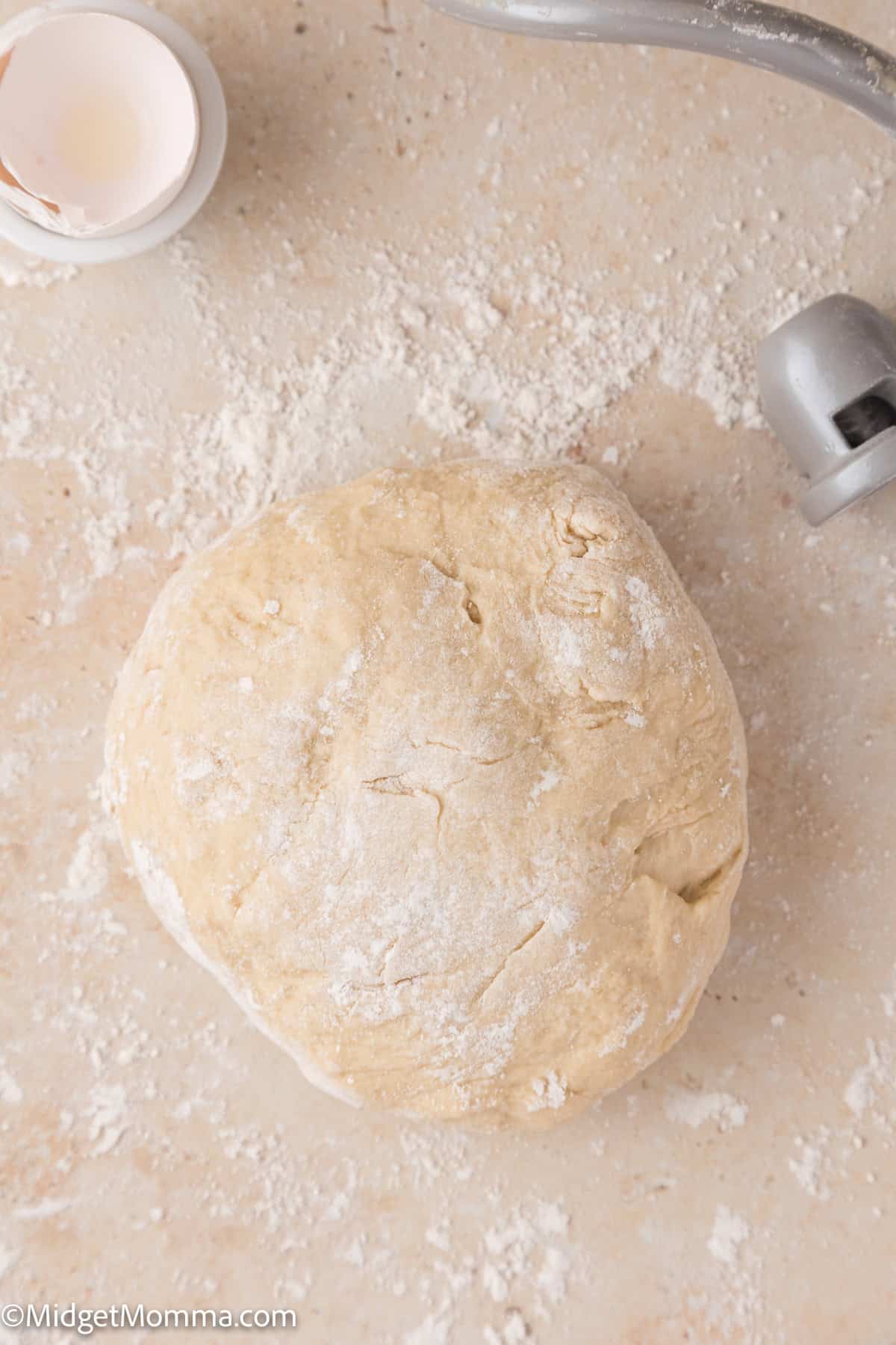 A ball of dough on a floured surface, with a small dish and a metal attachment nearby.