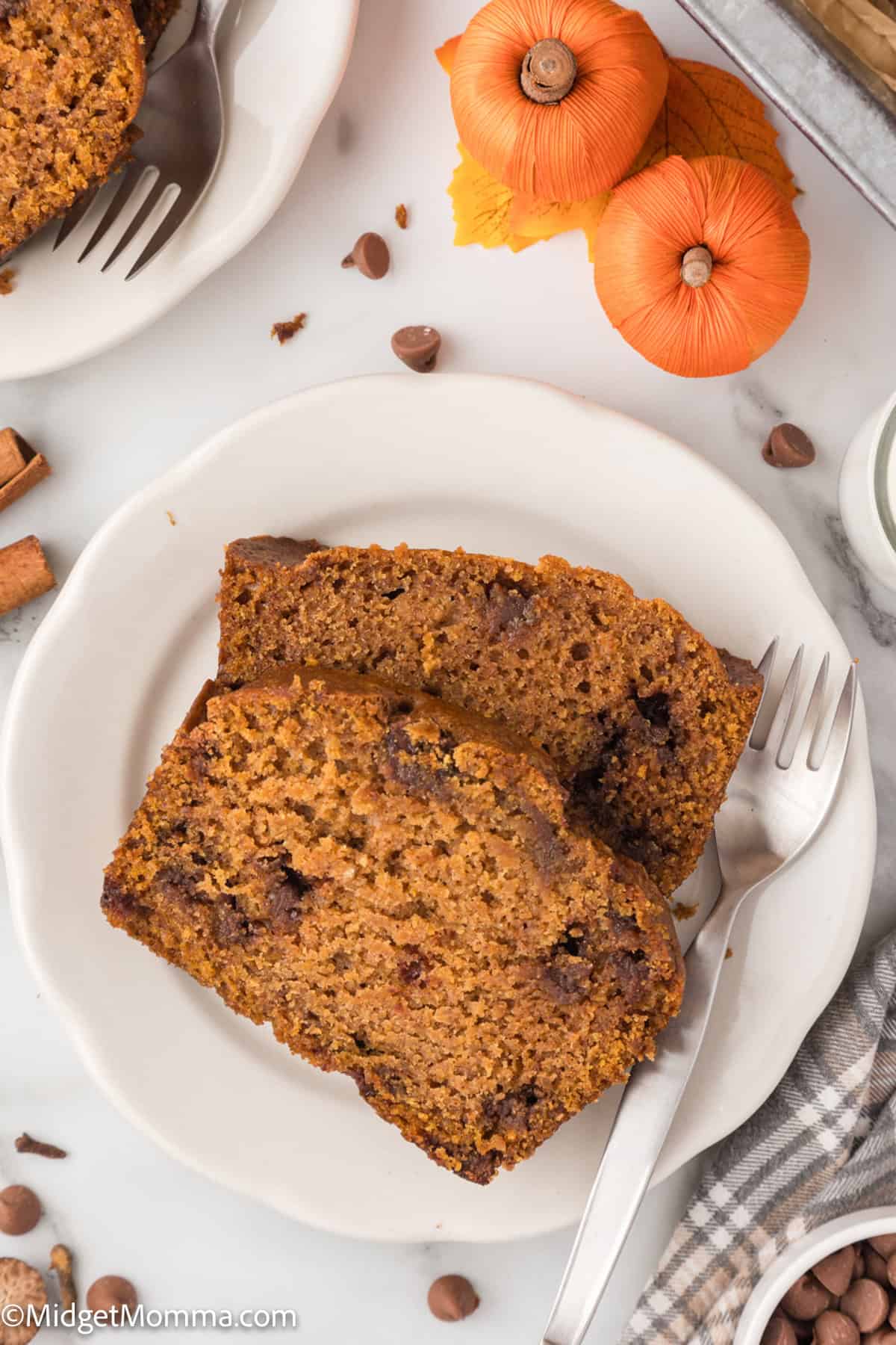 Two slices of pumpkin bread on a white plate with a fork, surrounded by mini pumpkins, cinnamon sticks, and chocolate chips on a marble surface.