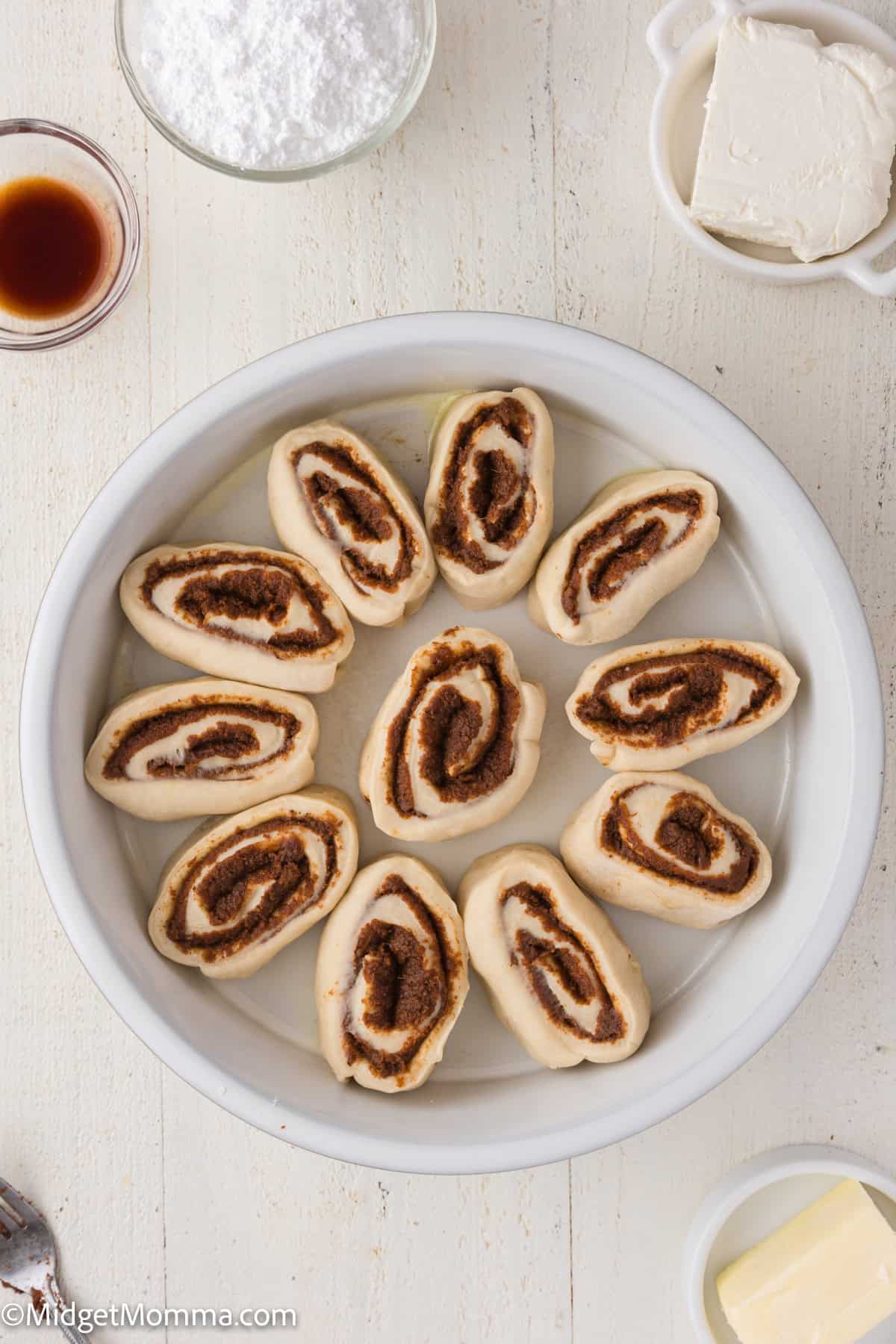 Unbaked cinnamon rolls arranged in a round baking dish. Ingredients like cream cheese, vanilla, butter, and powdered sugar are visible in the background.