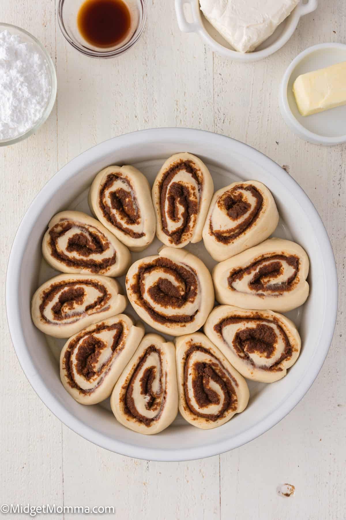 A circular arrangement of unbaked cinnamon rolls in a round baking dish on a white wooden surface. Ingredients like cream cheese, butter, and vanilla are nearby.