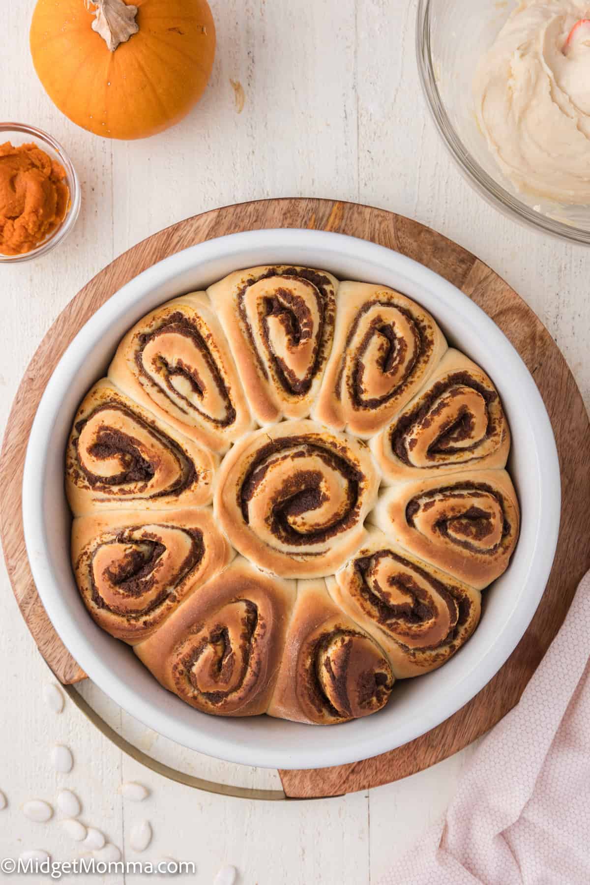 A round pan of spiral cinnamon rolls on a wooden board, with a small pumpkin and a bowl of frosting nearby.