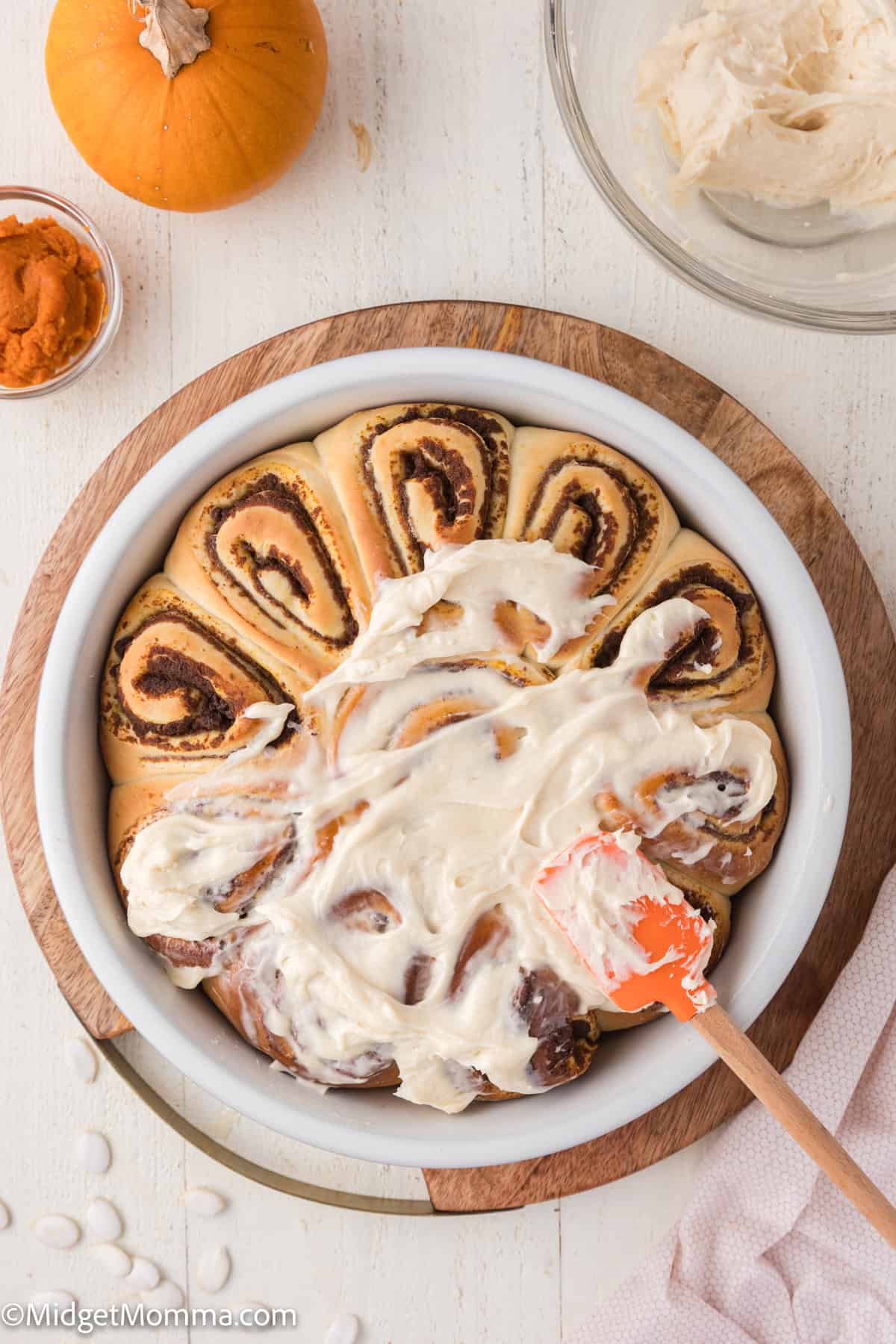 A round pan of cinnamon rolls partially covered with cream cheese frosting, next to a small pumpkin and a bowl of frosting.