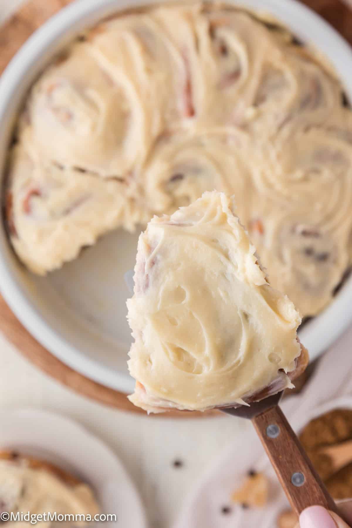 A close-up of a frosted cinnamon roll being held above a round baking dish filled with more frosted rolls.