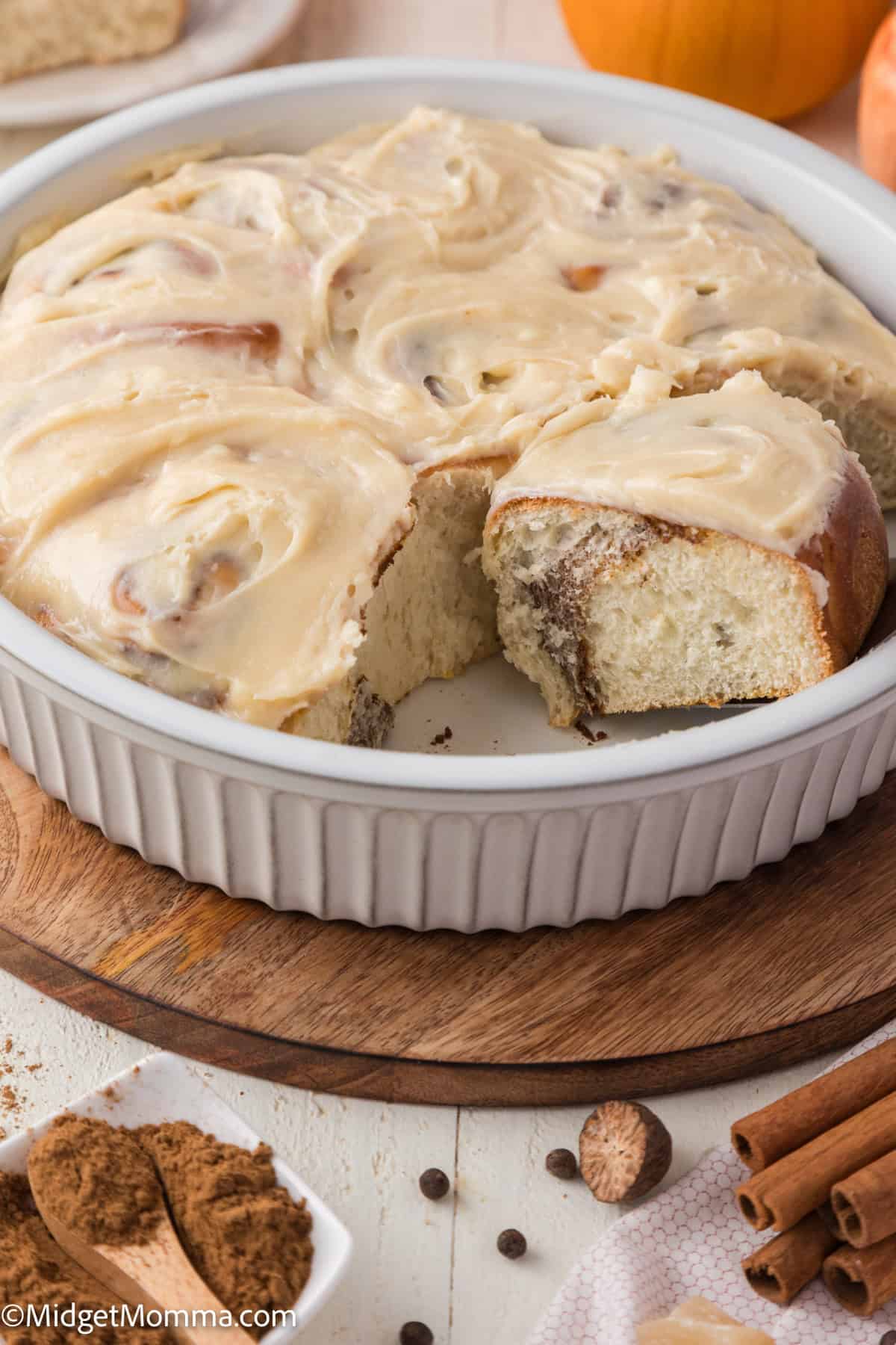 pumpkin cinnamon rolls with swirled frosting in a white dish with a slice missing. Cinnamon sticks and a small dish with powder are nearby on a wooden board.