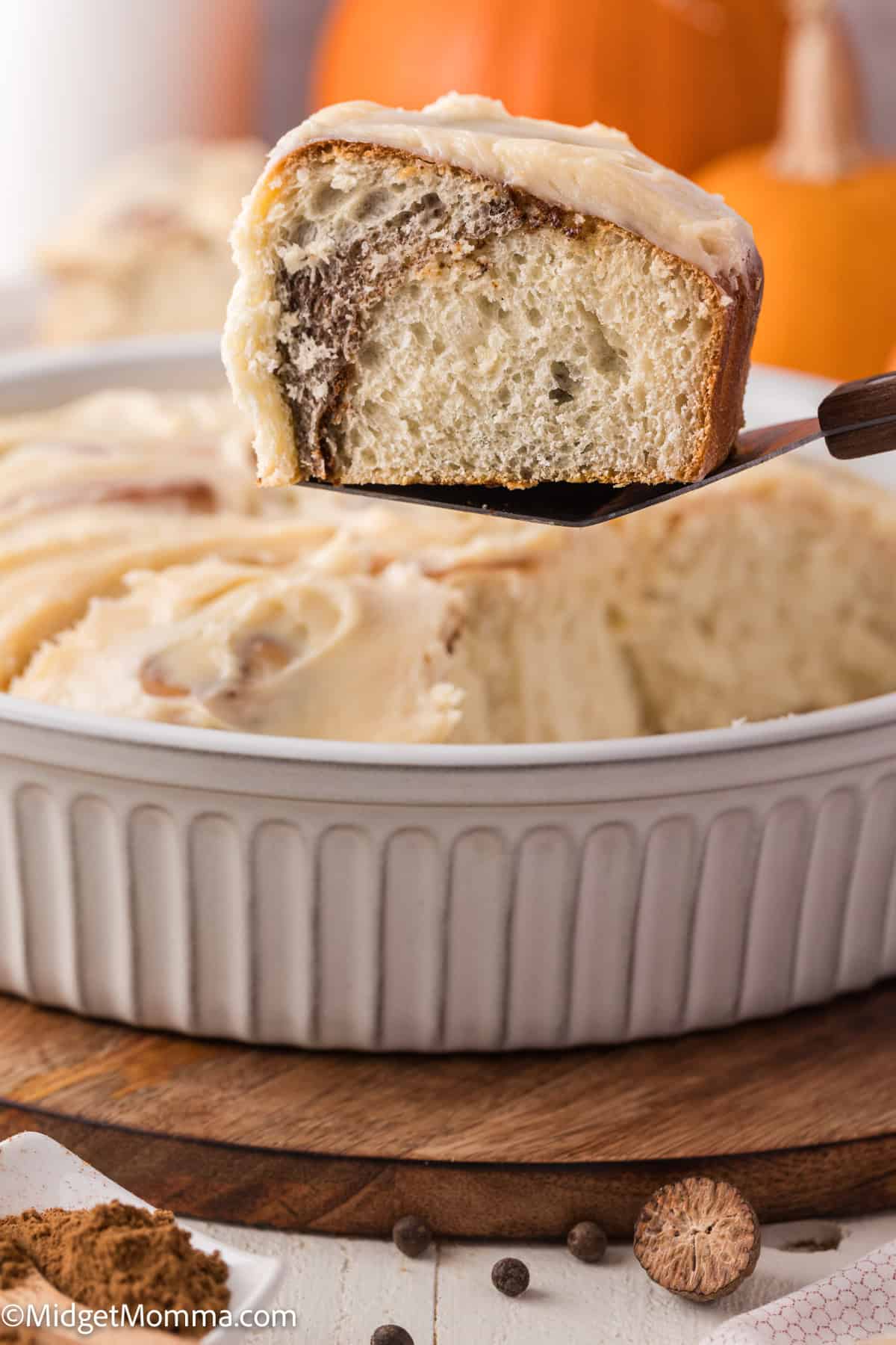 A close-up of a cinnamon roll with frosting, partially lifted from a round baking dish. A pumpkin is blurred in the background.