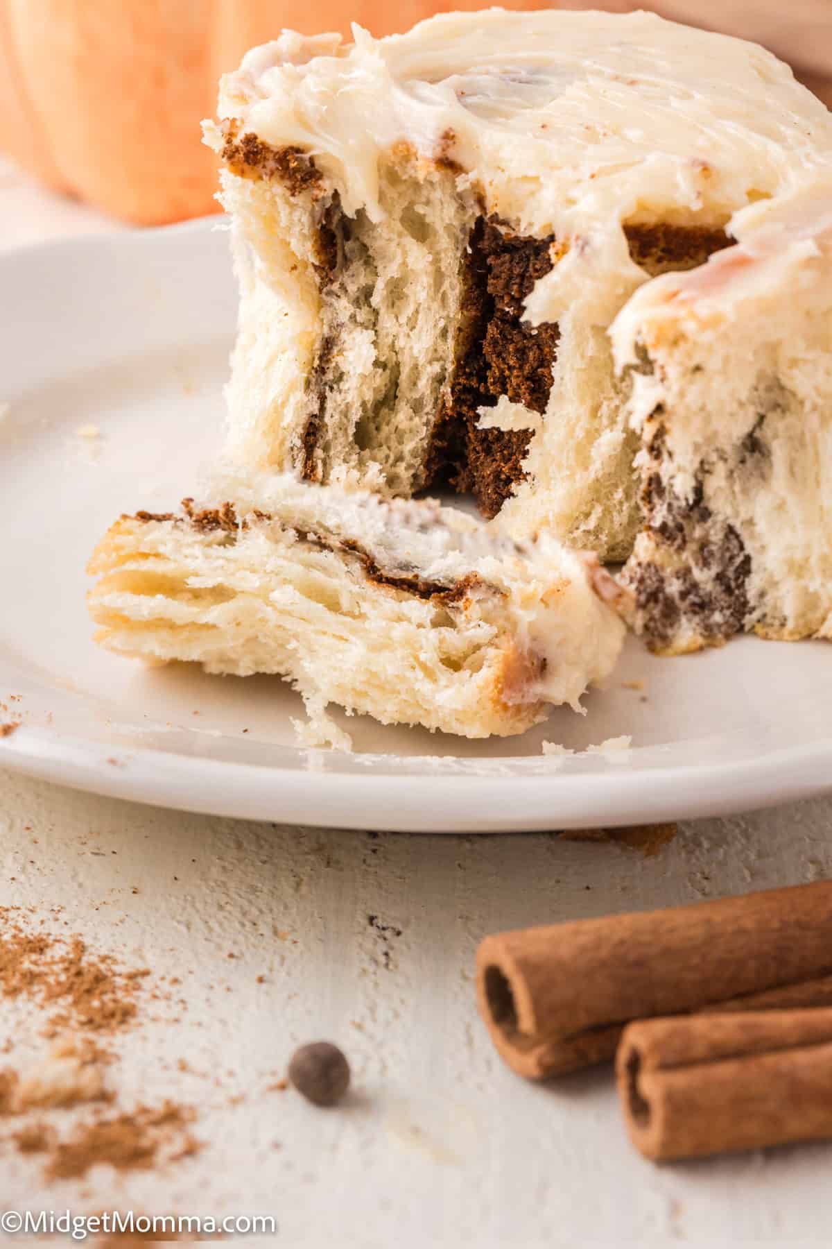 A cream cheese frosted cinnamon roll with a missing bite sits on a white plate, next to whole cinnamon sticks and scattered ground cinnamon.