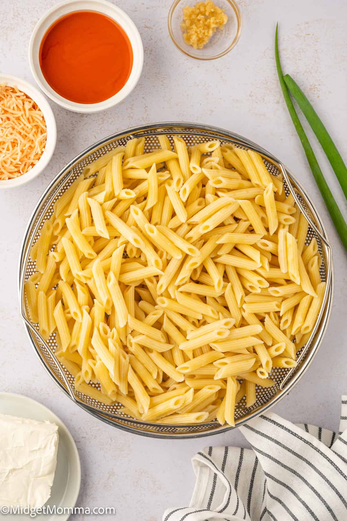 A bowl of cooked penne pasta surrounded by bowls of shredded cheese, cream cheese, minced garlic, and hot sauce. A striped cloth napkin and green onions are on the side.