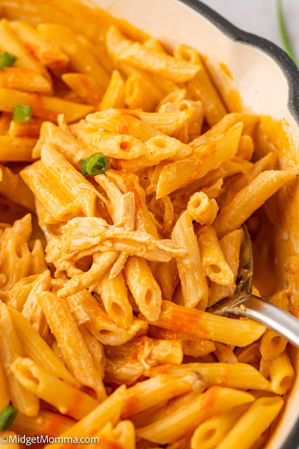 Close-up of a dish with penne pasta in a creamy buffalo sauce, garnished with green herbs. A spoon is partially visible, resting in the pasta.