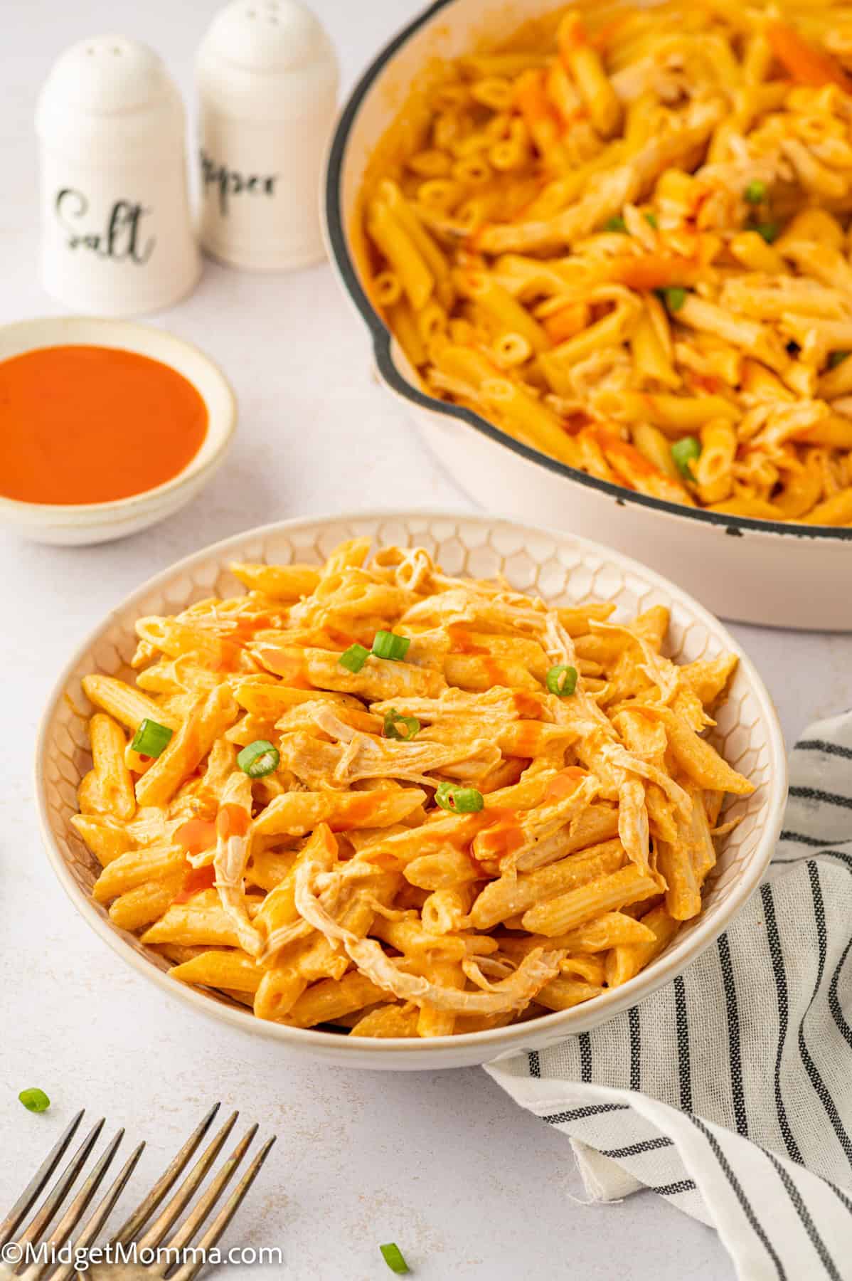 A bowl of creamy buffalo chicken pasta garnished with green onions. A serving dish of pasta, a small bowl of sauce, and salt and pepper shakers are in the background.