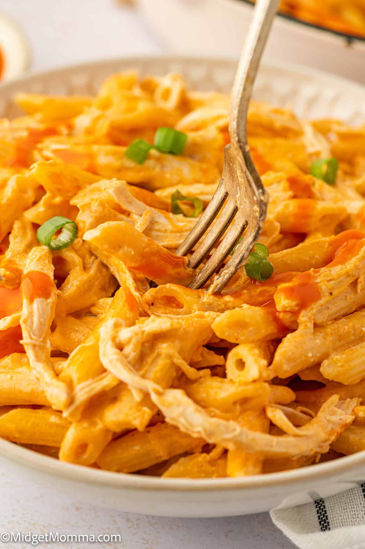 A fork picking up creamy penne pasta topped with shredded chicken, sliced green onions, and a drizzle of buffalo sauce on a white plate.