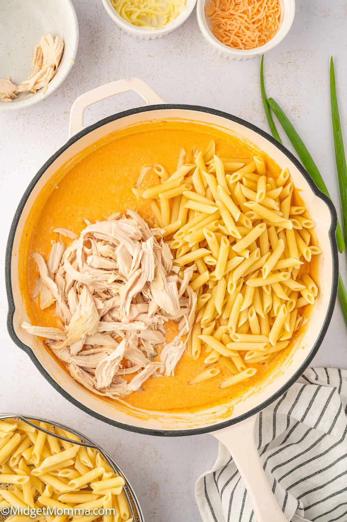 Pot of creamy buffalo sauce with shredded chicken and penne pasta on one side. Surrounding items include a towel, green onions, two small bowls with cheese, and a bowl with penne.