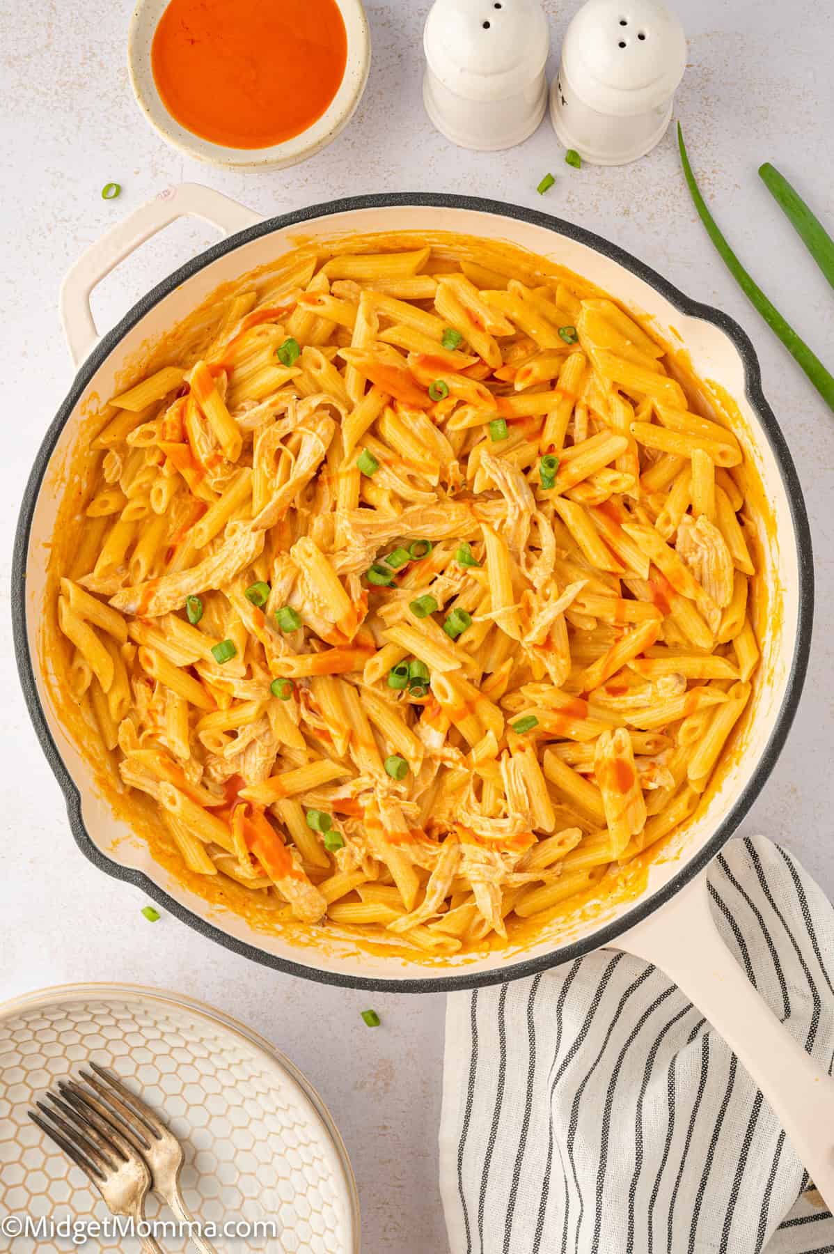 A pot of penne pasta with shredded chicken and vegetables, garnished with green onions, on a table alongside a striped napkin, plates, and a bowl of buffalo sauce.