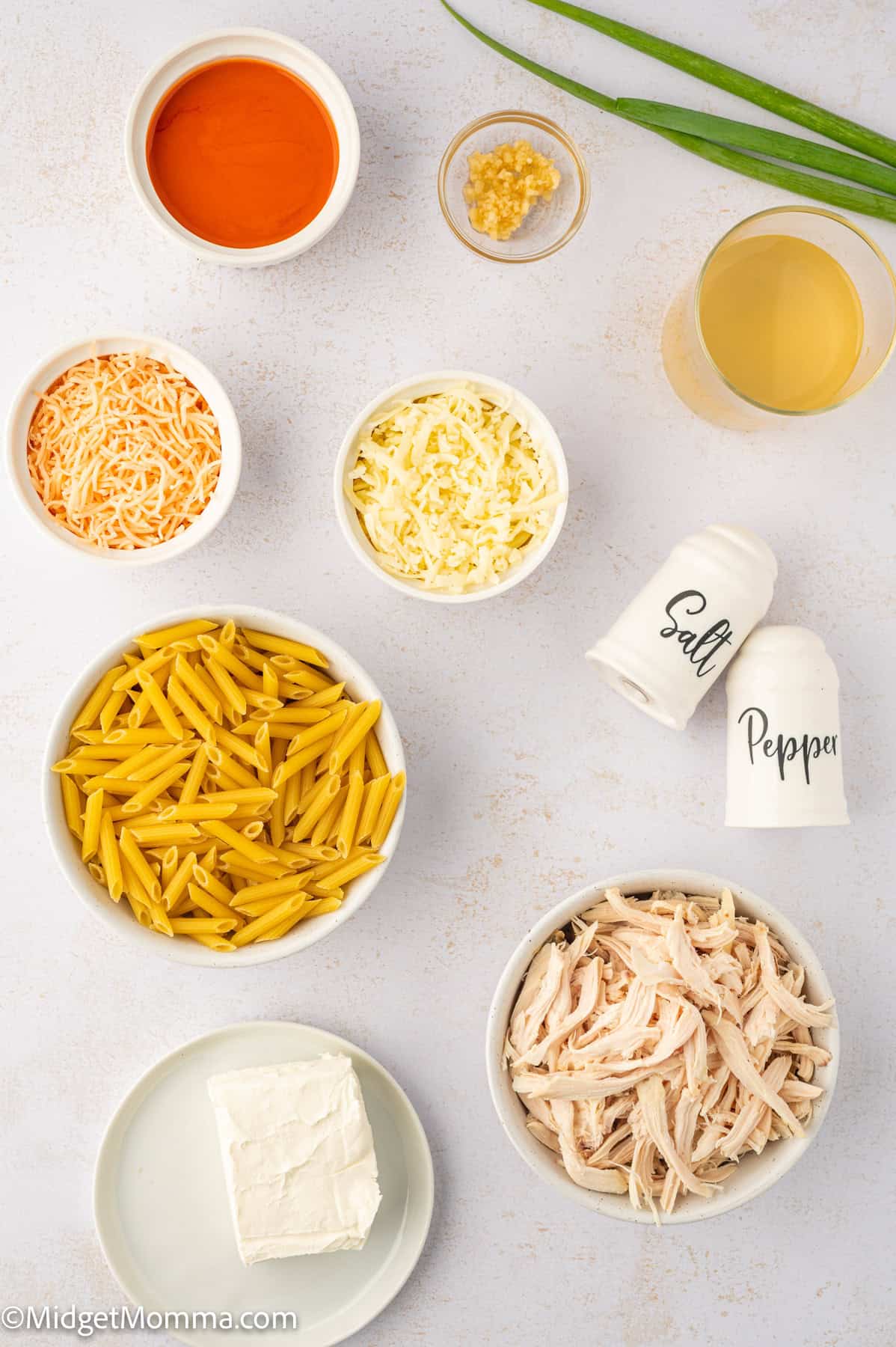 Buffalo Chicken Pasta Recipe ingredients. Ingredients for a dish including shredded chicken, pasta, shredded cheese, cream cheese, garlic, buffalo sauce, broth, and salt and pepper shakers. Scallions are visible in the background.