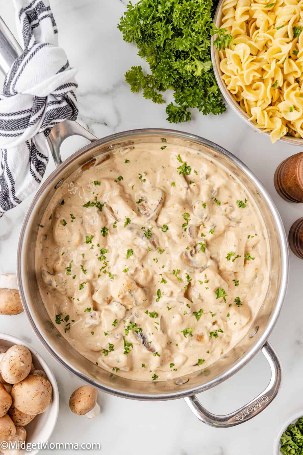 A pot of creamy chicken stroganoff mushroom sauce garnished with parsley, surrounded by raw mushrooms, parsley, and a bowl of cooked pasta.