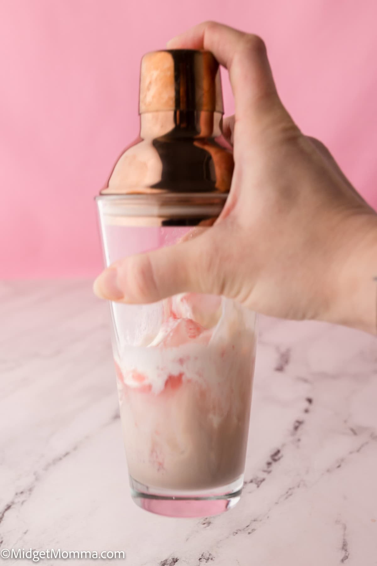 Hand holding a cocktail shaker filled with a creamy drink, against a pink background.