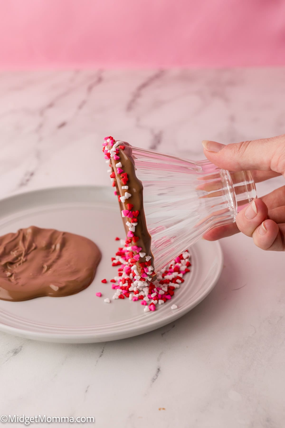 A hand dips the rim of an inverted glass into melted chocolate and sprinkles on a plate.