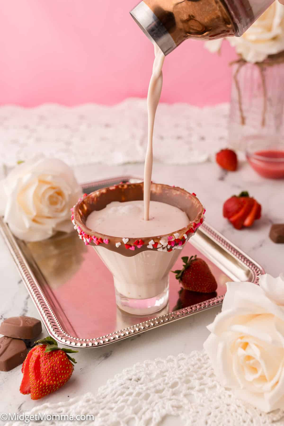 A cocktail is being poured into a rimmed glass on a tray, surrounded by white roses and strawberries.