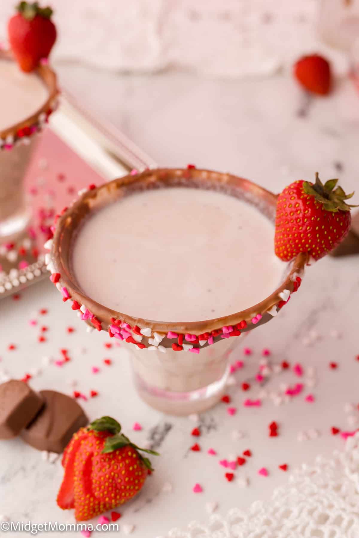 A pink cocktail drink in a glass with a chocolate-coated rim and heart-shaped sprinkles, garnished with a fresh strawberry.