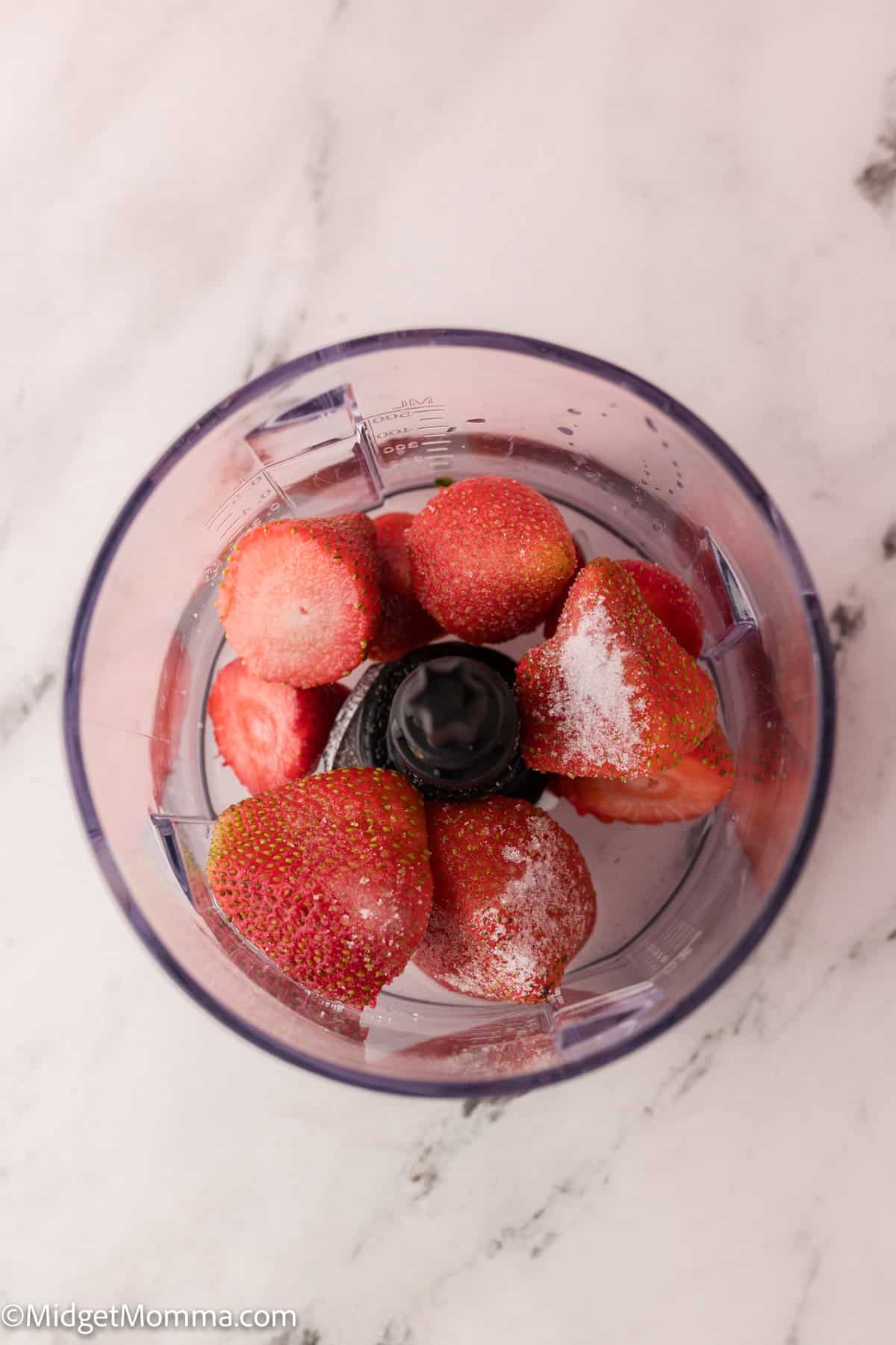 Top view of a blender filled with whole strawberries, some with frost, on a marble surface.