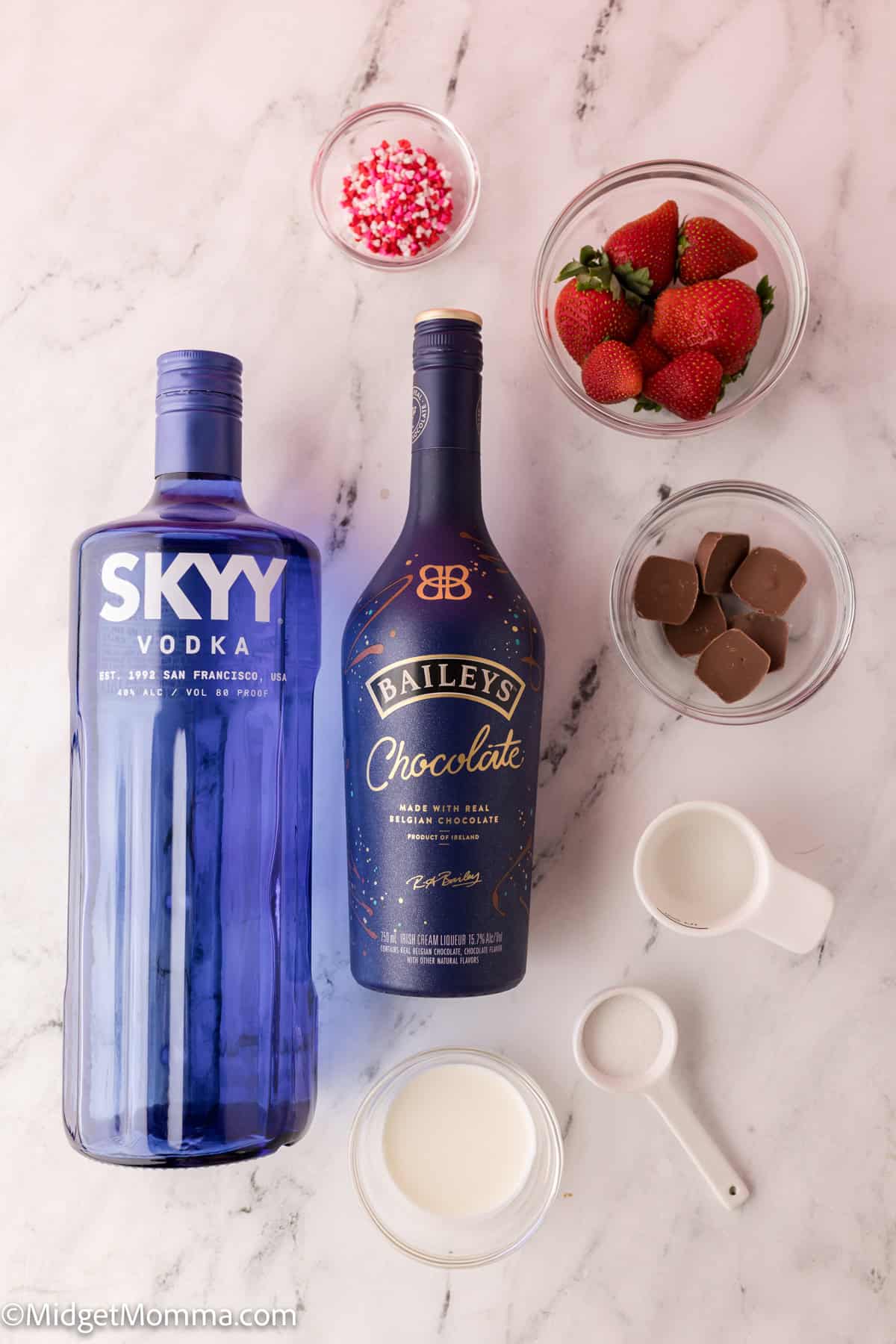 Chocolate Covered Strawberry Cocktail Recipe ingredients. Two bottles of Skyy Vodka and Baileys Chocolate on a marble surface, with bowls of strawberries, chocolate pieces, sprinkles, and measuring cups of cream.