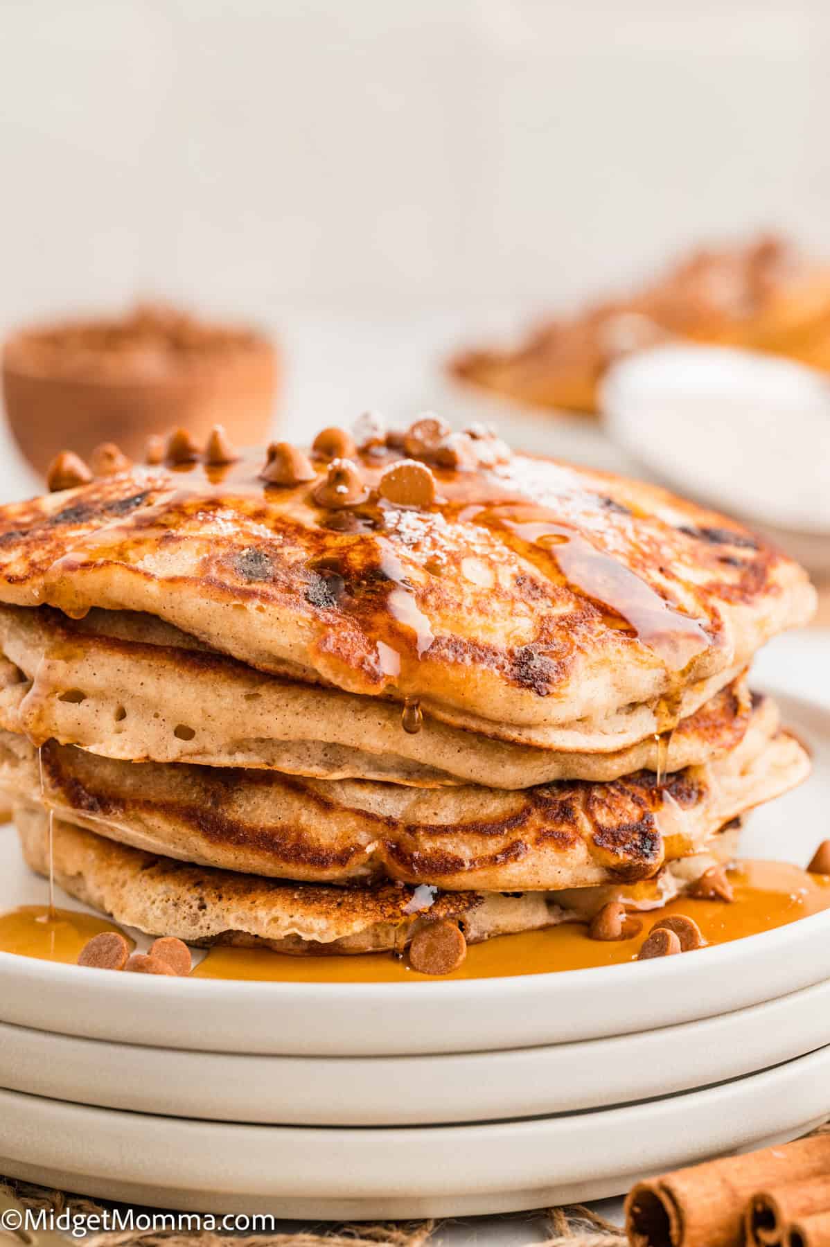 A stack of pancakes topped with syrup and cinnamon chips on a white plate. A bowl and cinnamon sticks are blurred in the background.