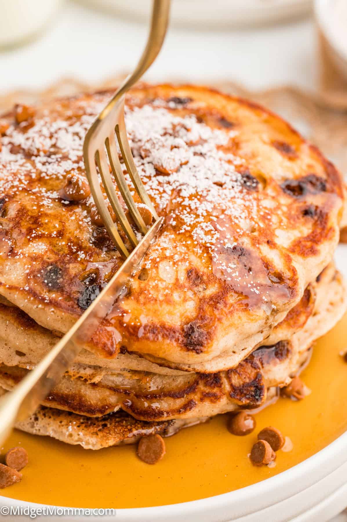 Stack of pancakes with cinnamon chips, dusted with powdered sugar, topped with syrup. A fork and knife are cutting into the stack.