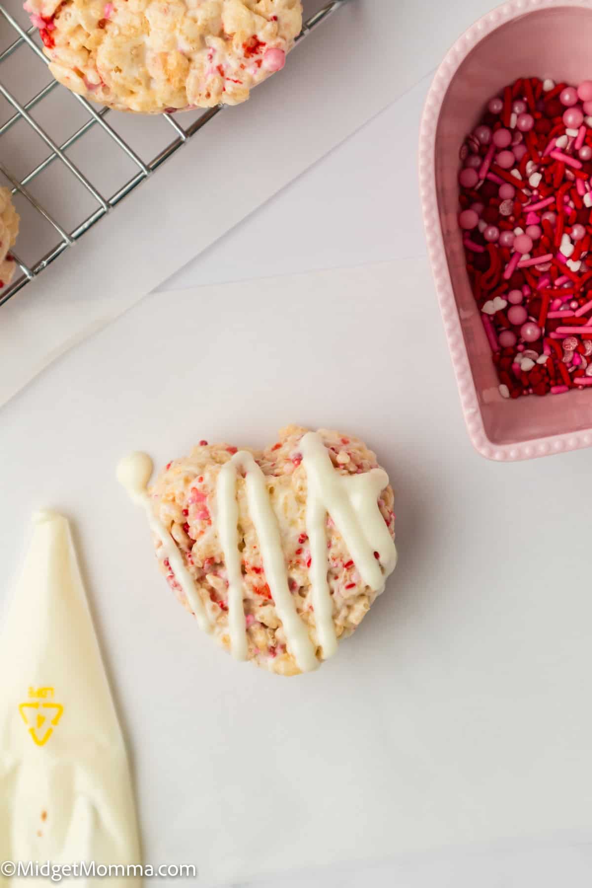 Heart-shaped Rice Krispie treat with white icing drizzle on parchment paper, pink bowl with red and pink sprinkles nearby, and a piping bag of icing.