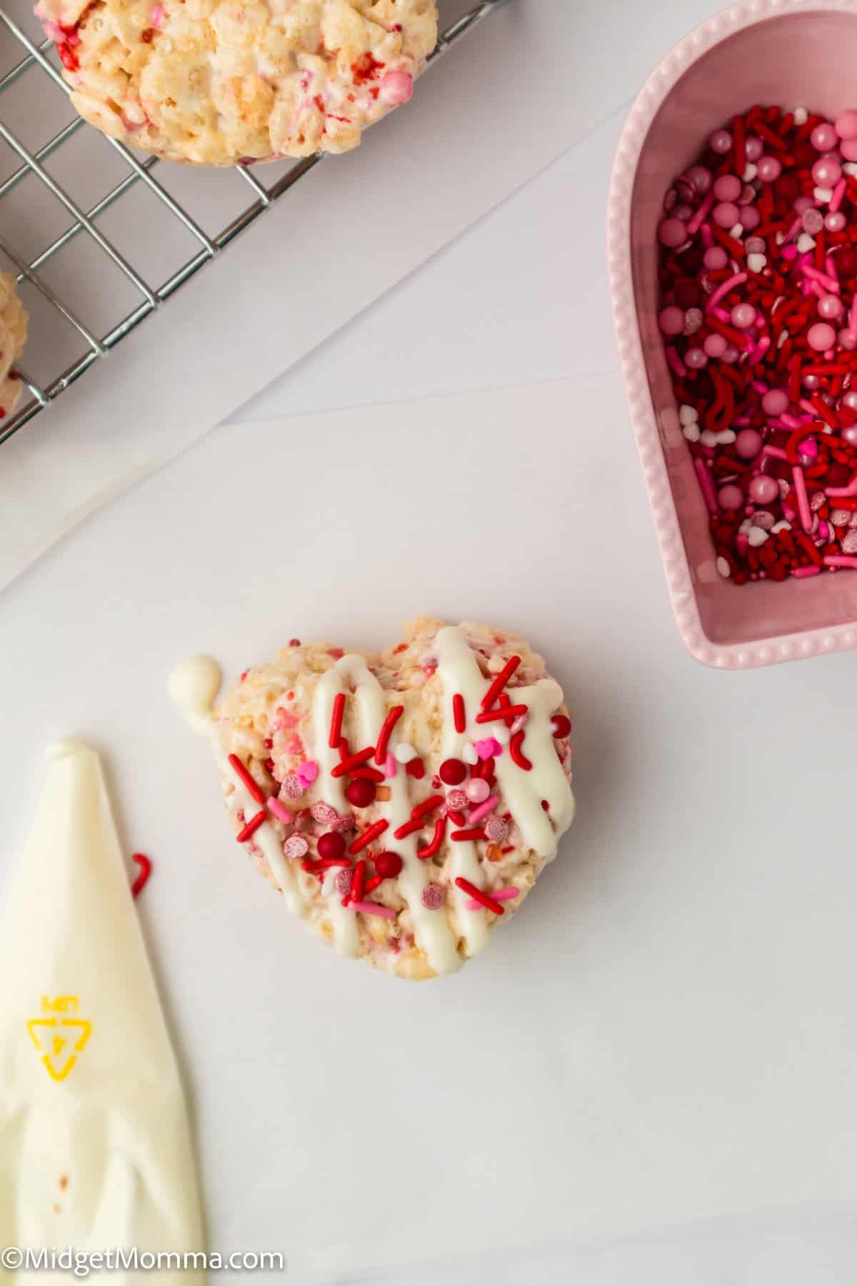 Heart-shaped Rice Krispie treat with white icing and red sprinkles beside a piping bag and pink container of sprinkles.