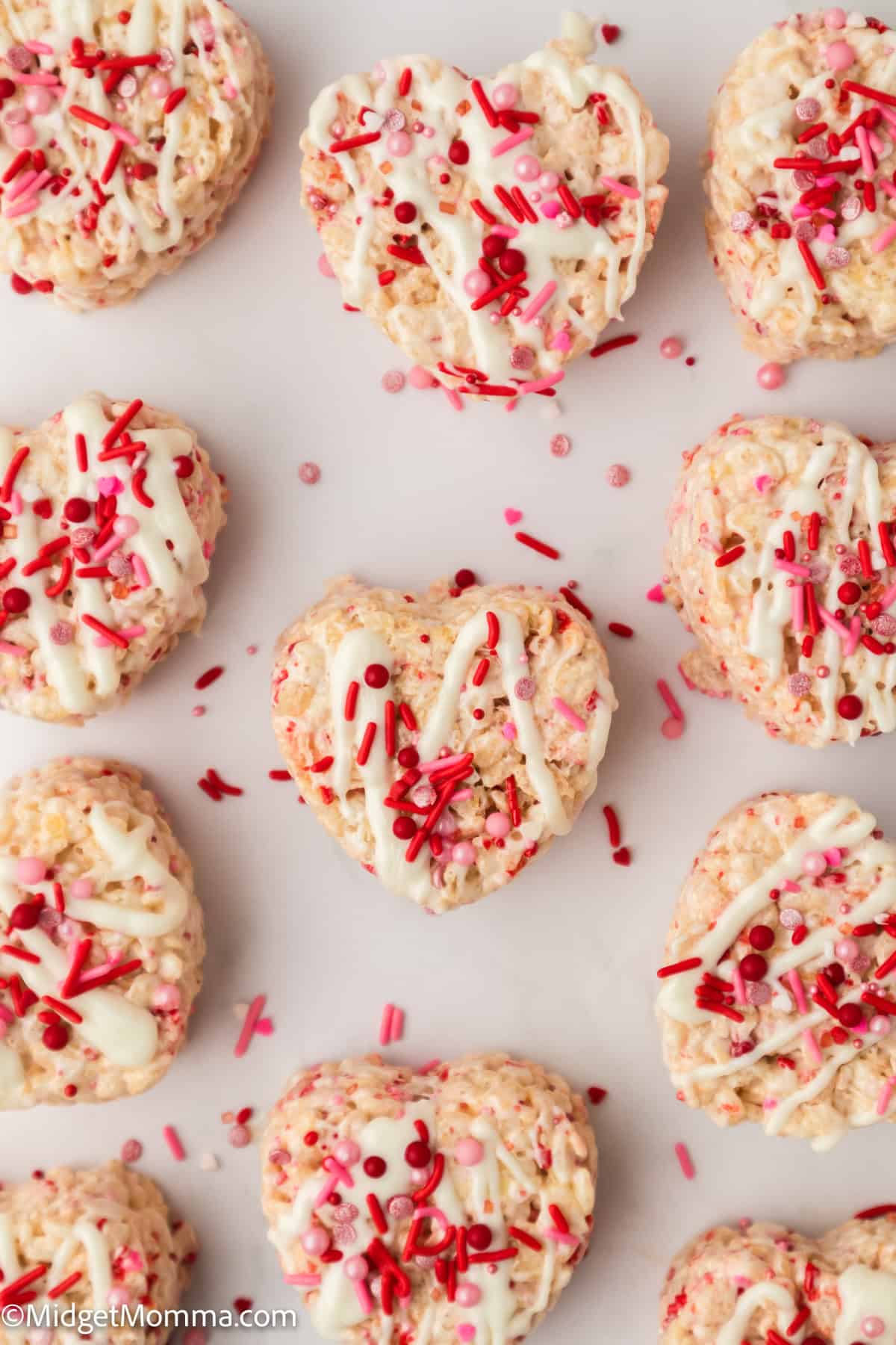 Heart-shaped Rice Krispie treats with white icing and red and pink sprinkles are arranged on a white surface.