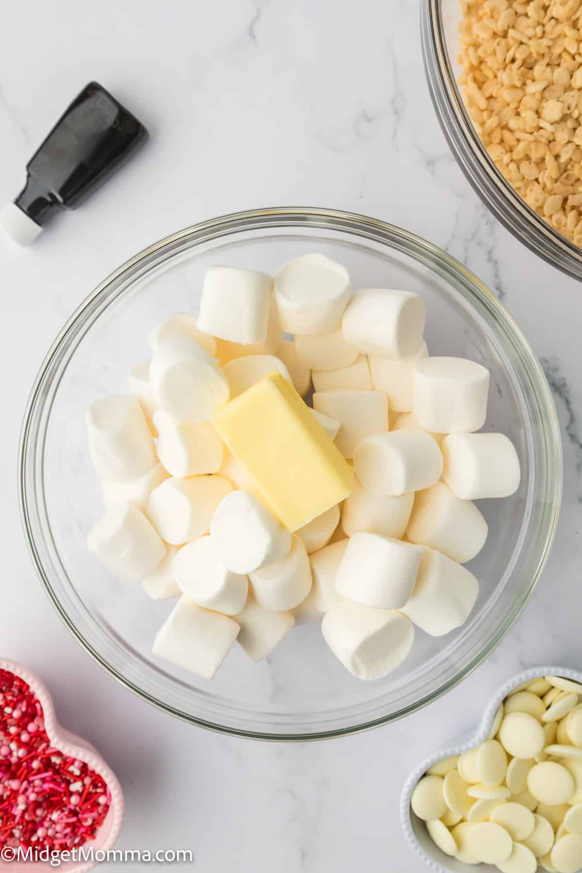 A bowl filled with marshmallows and a piece of butter is on a counter. Nearby are a bottle cap, a bowl of crisped rice, red sprinkles, and white chocolate chips.