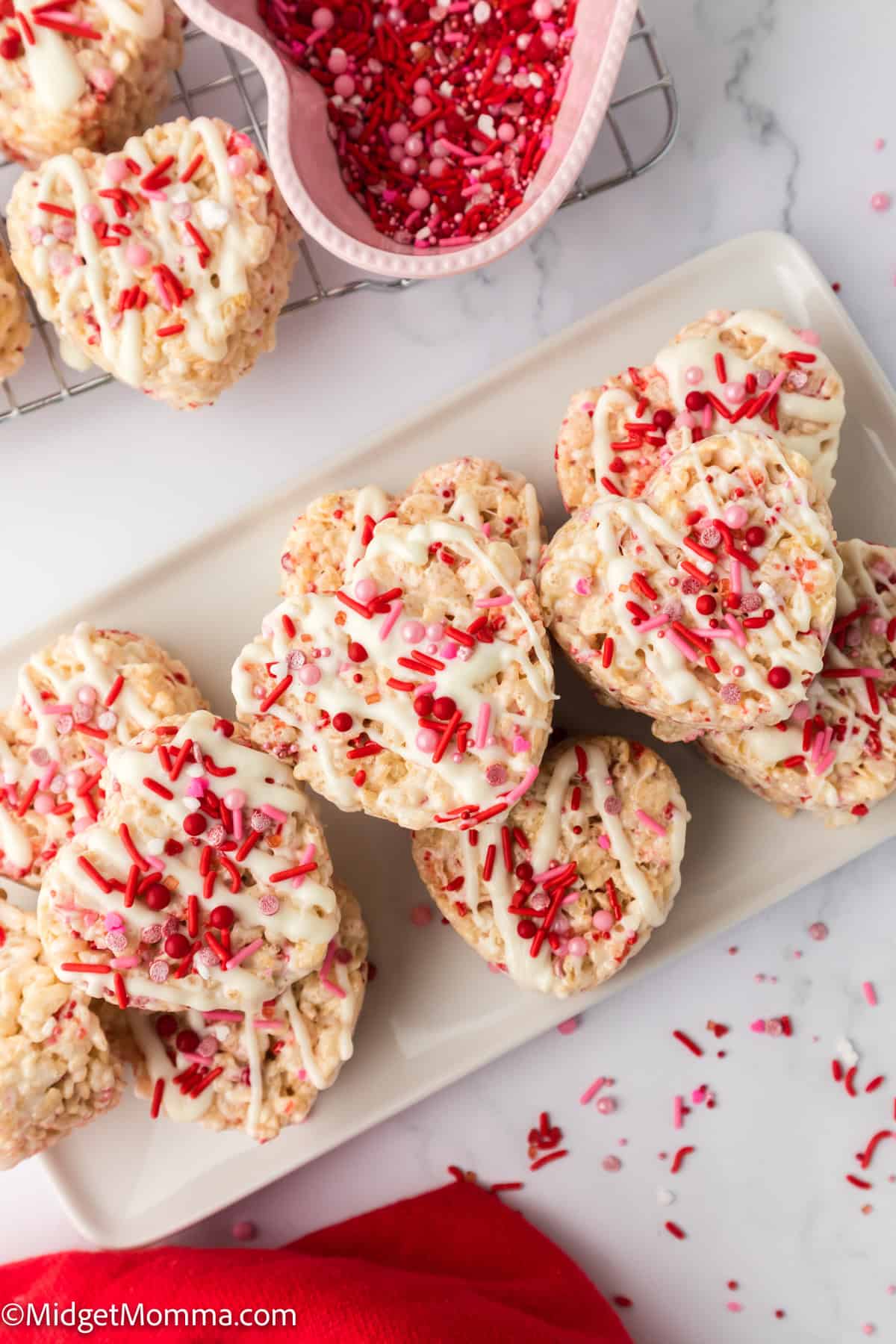 Heart-shaped Rice Krispie treats with white icing and red sprinkles on a white rectangular plate. Red sprinkles in a heart-shaped bowl and red cloth nearby.